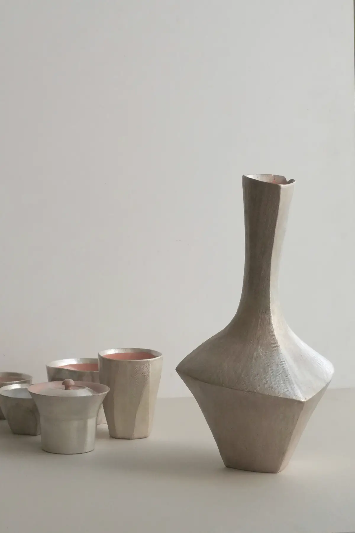 Bodegón de cerámica: un jarrón blanco facetado de cuello alto y varios vasos pequeños sobre fondo claro